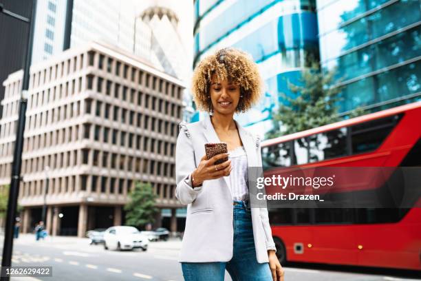 young curly woman using smart phone in downtown. double decker bus is in the background - double decker bus stock pictures, royalty-free photos & images