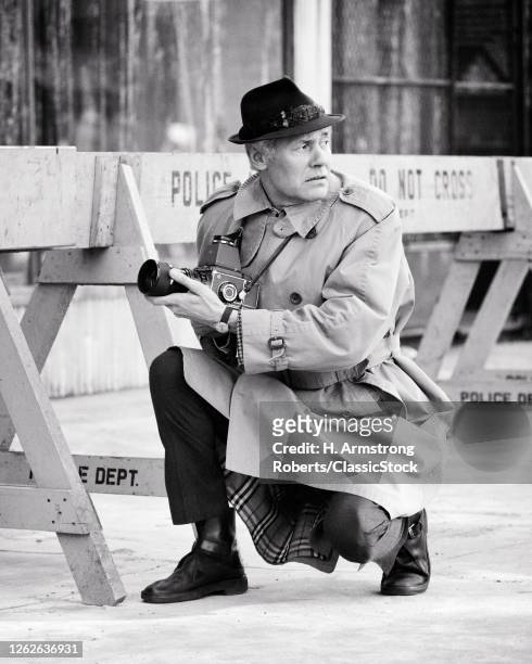 1970s Man Professional Photographer Wearing Trench Coat And Snap Brim Hat Kneeling By Police Barrier Looking Back Over Shoulder