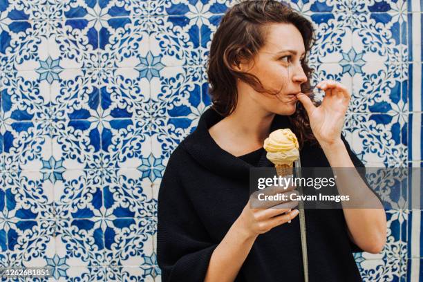 millennial woman licking finger while eating ice cream at a tiled wall - frau eistüte stock-fotos und bilder