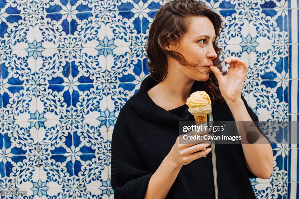 Millennial woman licking finger while eating ice cream at a tiled wall