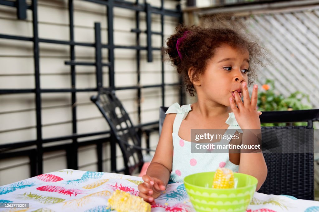 Mixed-race toddler eating corn outdoors in summer.