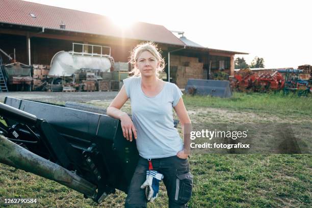 portrait: female farmer leans against a tractor, looking at camera - white barn stock pictures, royalty-free photos & images