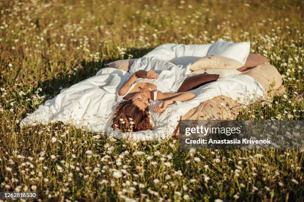 young cute curly woman in white dress lying on soft linen bed in summer chamomile field. summer vacations inspiration. concept of good sleep and relax. - frau blumenwiese stock-fotos und bilder