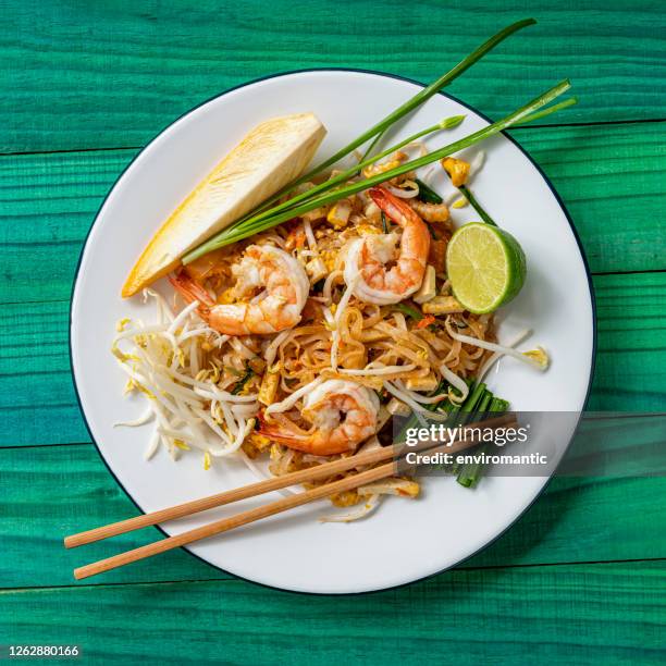 ricetta thailandese di noodles thai di gamberi appena saltati in padella su un piatto rotondo tradizionale in metallo smaltato con bacchette posate sul lato del piatto, incastonato su uno sfondo astratto in legno color turchese. - noodle foto e immagini stock