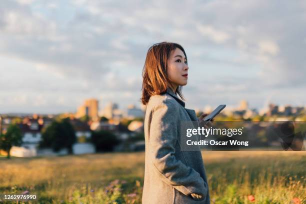 young woman holding a smart phone, watching sunrise at the park - asian woman smiling sunrise stock pictures, royalty-free photos & images