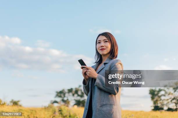 young woman holding a smart phone, watching sunrise at the park - asian woman smiling sunrise stock pictures, royalty-free photos & images