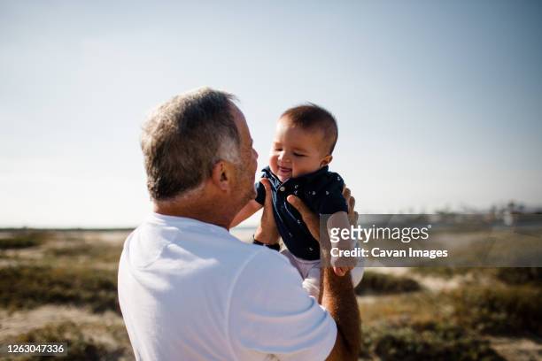 grandfather holding grandson while standing on beach - baby grandpa imagens e fotografias de stock