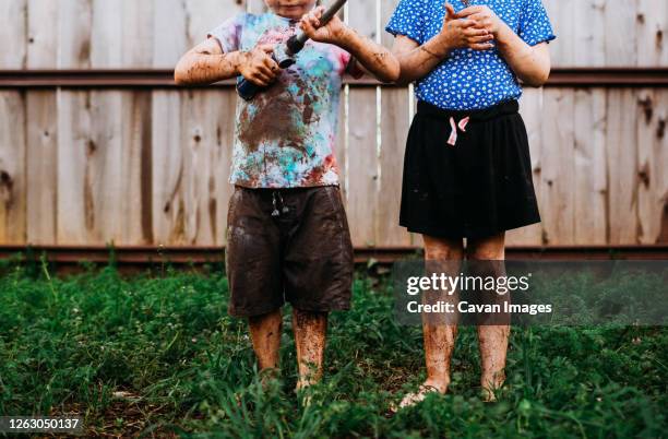 young brother and sister standing outside covered in mud - mud stock-fotos und bilder