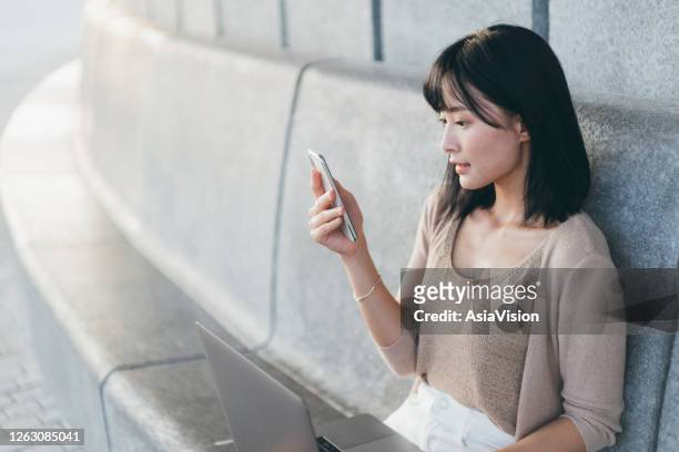 young asian woman using smartphone while working on a laptop outdoors in urban park - chinese student laptop stock pictures, royalty-free photos & images