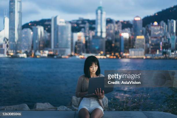 年輕的亞洲婦女使用筆記型電腦在維多利亞港的長廊,對照亮香港城市景觀在黃昏。 - 遊園地 個照片及圖片檔