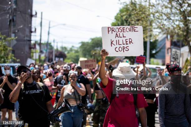 Protester wearing a mask and a hat holds a sign that says, "Prosecute All Killer Cops" in front of the crowd of protesters as they walk through...