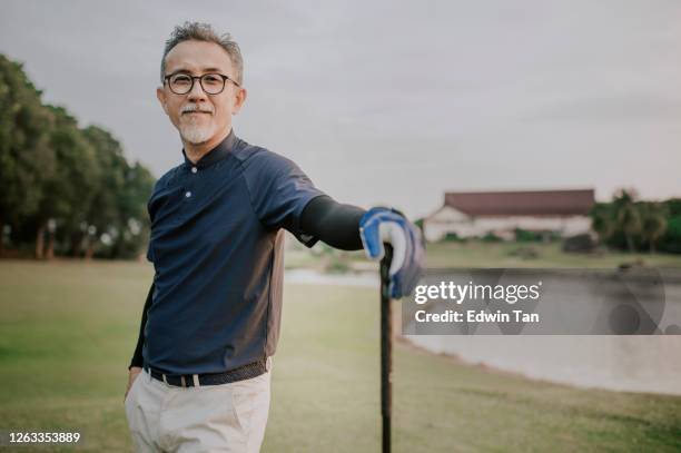 un asiático chino activo senior hombre golfista de pie con su conductor de club de golf en el campo de golf después del juego de golf esperando y buscando - golf sport fotografías e imágenes de stock