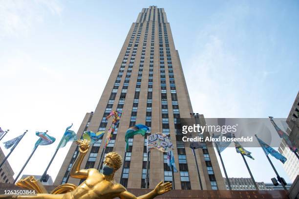 Flags that are part of "The Flag Project" are seen flying behind Paul Manship's 'Prometheus' statue wearing a mask with a view of the 30 Rockefeller...