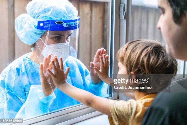 family reunion in times of pandemic: man holding his son looking at her healthcare worker mother through a window, child and father are indoors, mother outdoors - looking through window covid stock pictures, royalty-free photos & images