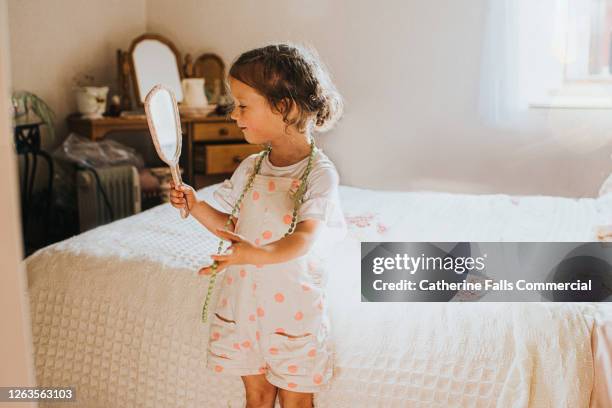 cute little girl in a dated bedroom tries on costume jewellery and looks into an old-fashioned hand mirror - modeschmuck stock-fotos und bilder