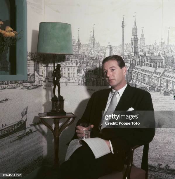 British dramatist and playwright Terence Rattigan posed holding a glass and a play script in a living room in 1952. The wall behind is decorated with...