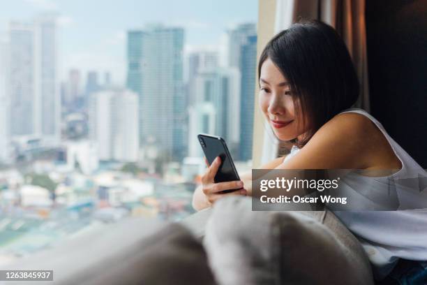 young woman checking social media on smart phone near the window - center for asian american media stock pictures, royalty-free photos & images