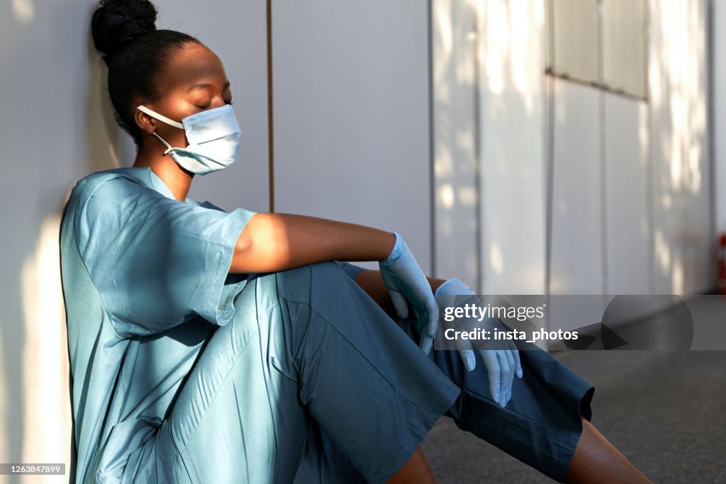 Tired exhausted female african scrub nurse wears face mask blue uniform gloves sits on hospital floor. Depressed sad black ethic doctor feels fatigue burnout stress, lack of sleep, napping at work.