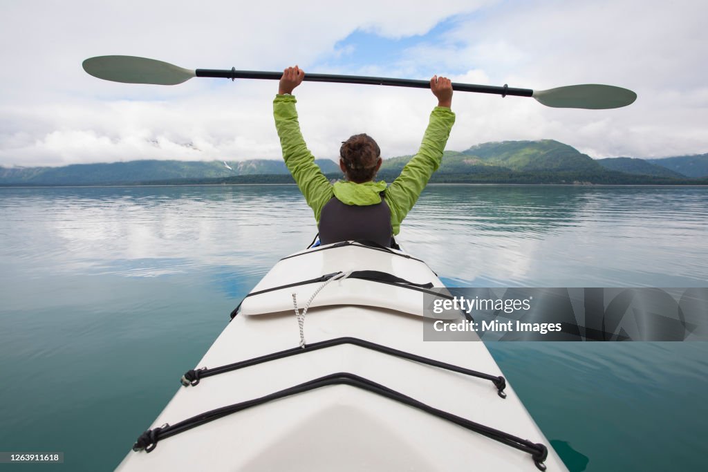 Sea kayaker extends paddle over her head, pristine waters of Muir Inlet in distance, Glacier Bay National Park, Alaska