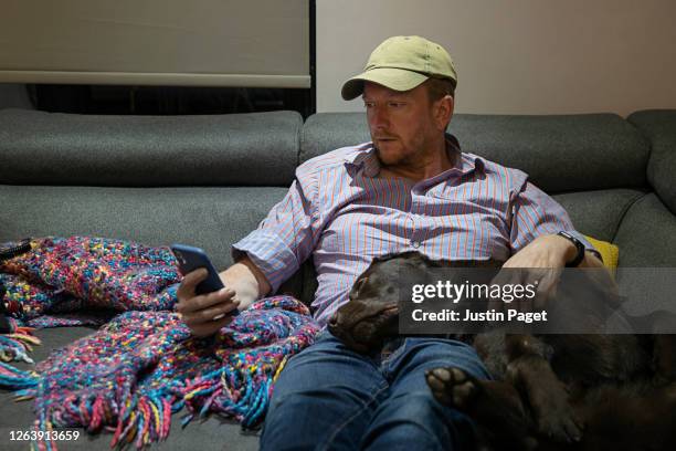 man on sofa with his pet dog. he surfs the net on his smartphone - man sleeping with cap stock pictures, royalty-free photos & images