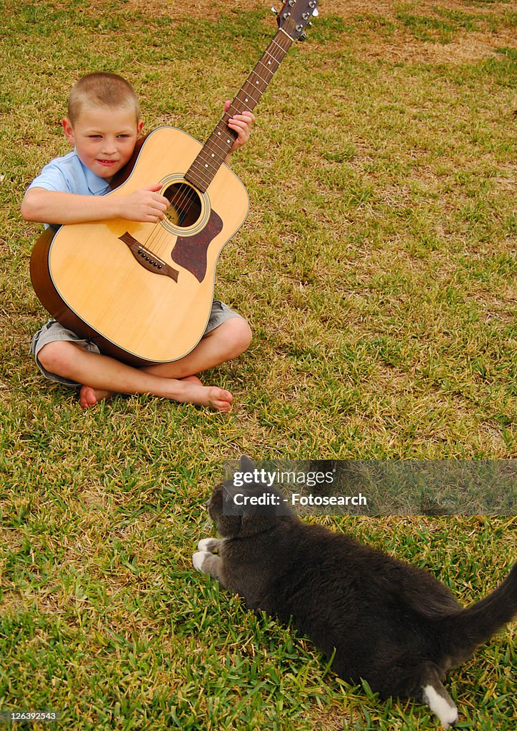 Boy playing a guitar with a cat sitting in front of him