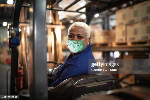 porträt einer arbeiterin, die gabelstapler im lager fährt - mit gesichtsmaske - essenzielle berufe und dienstleistungen stock-fotos und bilder