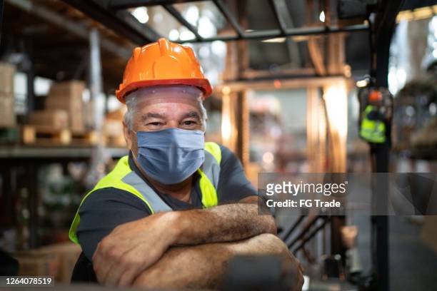 porträt eines älteren männlichen arbeiters, der gabelstapler in lagerhalle fährt - mit gesichtsmaske - essenzielle berufe und dienstleistungen stock-fotos und bilder
