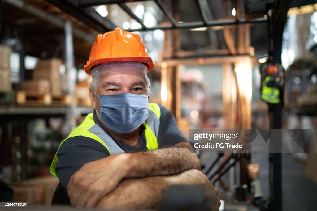 Portrait of senior male worker driving forklift in warehouse - using face mask