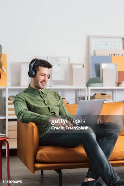 moderne arbeitsstationen: ein lächelnder freelancer, der im büro sitzt und kopfhörer aufundhört und musik hört, während er an seinem laptop-pc arbeitet - grünes hemd stock-fotos und bilder