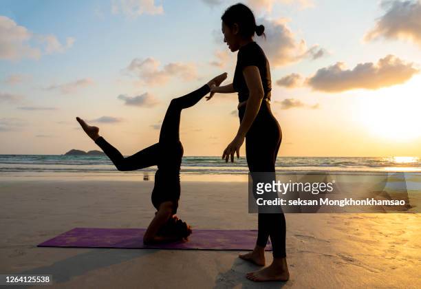 a woman practices yoga on the beach whilst another woman helps her. - yoga instructor stock pictures, royalty-free photos & images