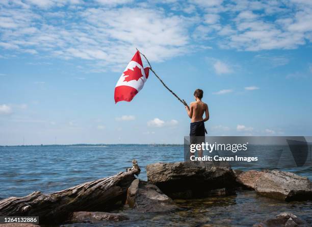 boy holding canada flag in the air on shore of a lake on a summer day. - canada day celebration stock pictures, royalty-free photos & images