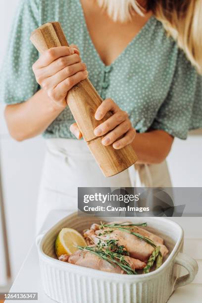 caucasian woman with a pepper grinder seasoning raw chicken with herbs - vleesmolen stockfoto's en -beelden