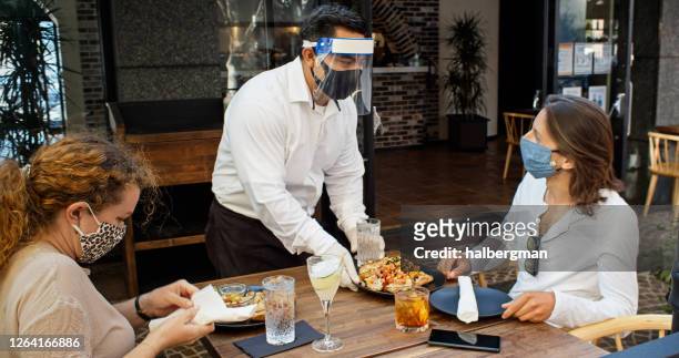 cameriere che indossa dpi durante la pandemia di covid-19 che serve cibo ai commensali indossando maschere - ristorante foto e immagini stock