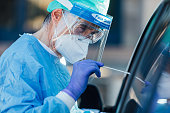 Medical personnel wearing a PPE, performing PCR with a swab in their hand, on a patient inside his car to detect if he is infected with COVID-19