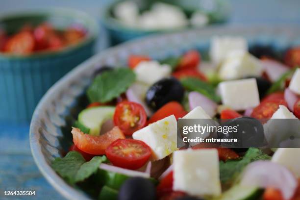 bild von griechischen (horiatiki) salat gemusterte schüssel, beilagen von gehackten kirschtomaten und feta-käse, gemüse einschließlich, tomaten, gurken, zwiebel, feta-käse, minzblätter und schwarze oliven, blauer holzhintergrund, fokus auf den vorder - griechischer salat stock-fotos und bilder