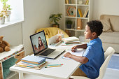 Focused black schoolboy with curly hair sitting at desk and using laptop while listening to tutor during video conference