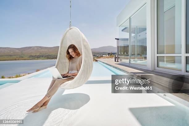 woman sitting in hanging chair above swimming pool using laptop - design laptop woman stock-fotos und bilder