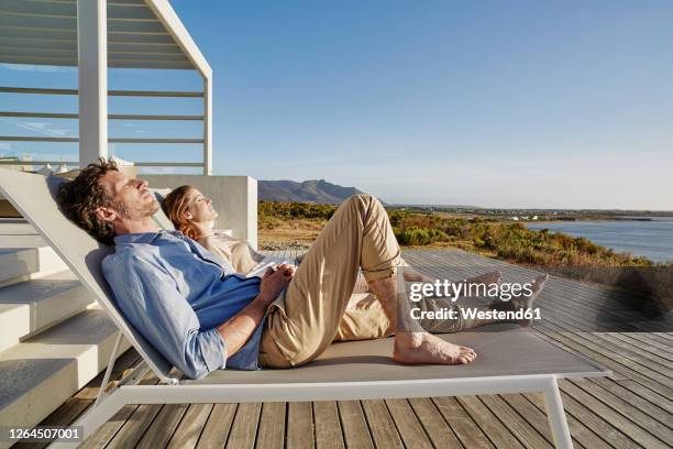 couple lying on deck chairs at luxury beach house - sedia a sdraio foto e immagini stock