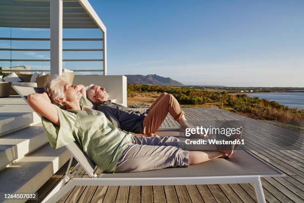 senior couple lying on deck chairs at luxury beach house - casa al mare foto e immagini stock