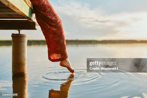 woman sitting on jetty at a lake at sunset touching the water with her foot - legs in water stock pictures, royalty-free photos & images