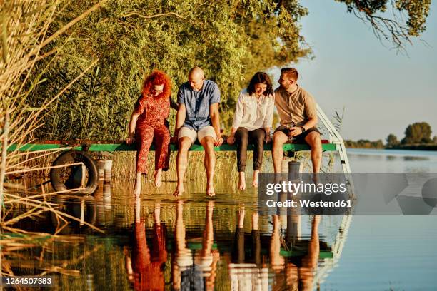friends reflected in water sitting on jetty at a lake - four people foto e immagini stock