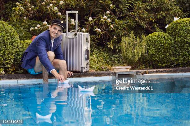 smiling man floating paper boats on swimming pool while sitting by suitcase against plants - spielzeugschiff stock-fotos und bilder