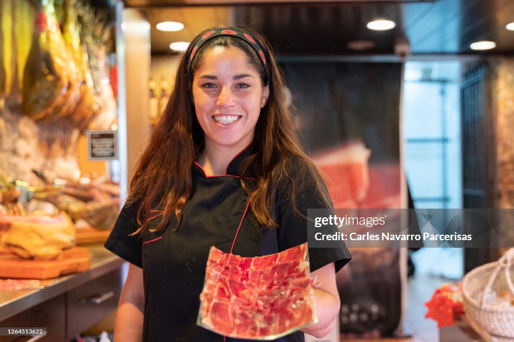 Woman in uniform showing a paper full of ham slices while is facing the camera on a butcher