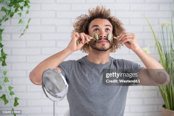 young man massaging face with jade roller against wall at home - 玉石滾輪 個照片及圖片檔