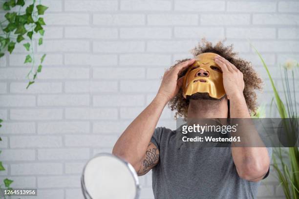 young man applying facial mask while sitting against wall at home - mask man stock pictures, royalty-free photos & images