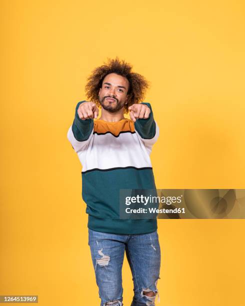 handsome young man with curly hair pointing while standing against yellow background - ripped jeans stock pictures, royalty-free photos & images