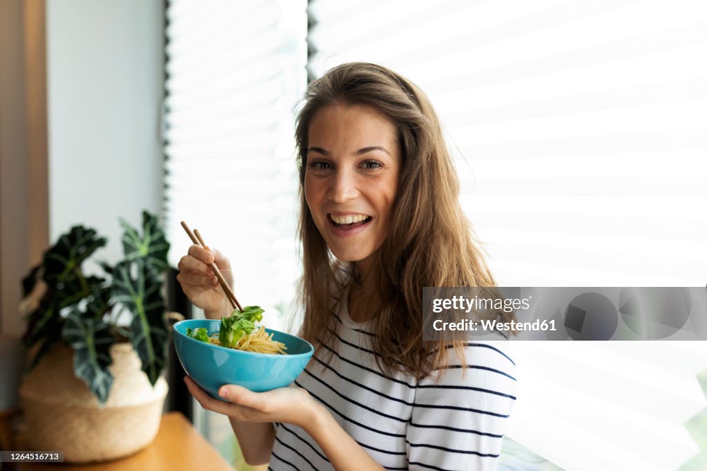 Happy young woman holding food and chopsticks while standing against window at home