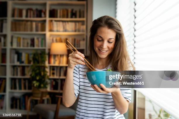 smiling young woman eating food while sitting by window at home - chopsticks stock-fotos und bilder