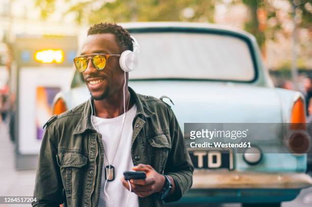 smiling young man wearing sunglasses listening music through headphones against vintage car in city - music from the motor city stock pictures, royalty-free photos & images
