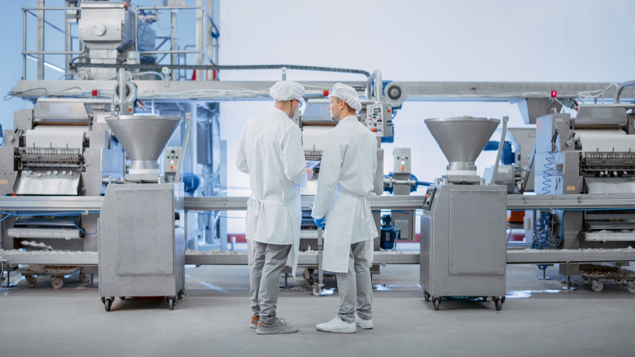 Two Young Food Factory Employees Discuss Work-Related Matters. Male Technician or Quality Manager Uses a Tablet Computer for Work. They Wear White Sanitary Hat and Work Robes. Two Young Food Factory Employees Discuss Work-Related Matters. Male Technician or Quality Manager Uses a Tablet Computer for Work. They Wear White Sanitary Hat and Work Robes.